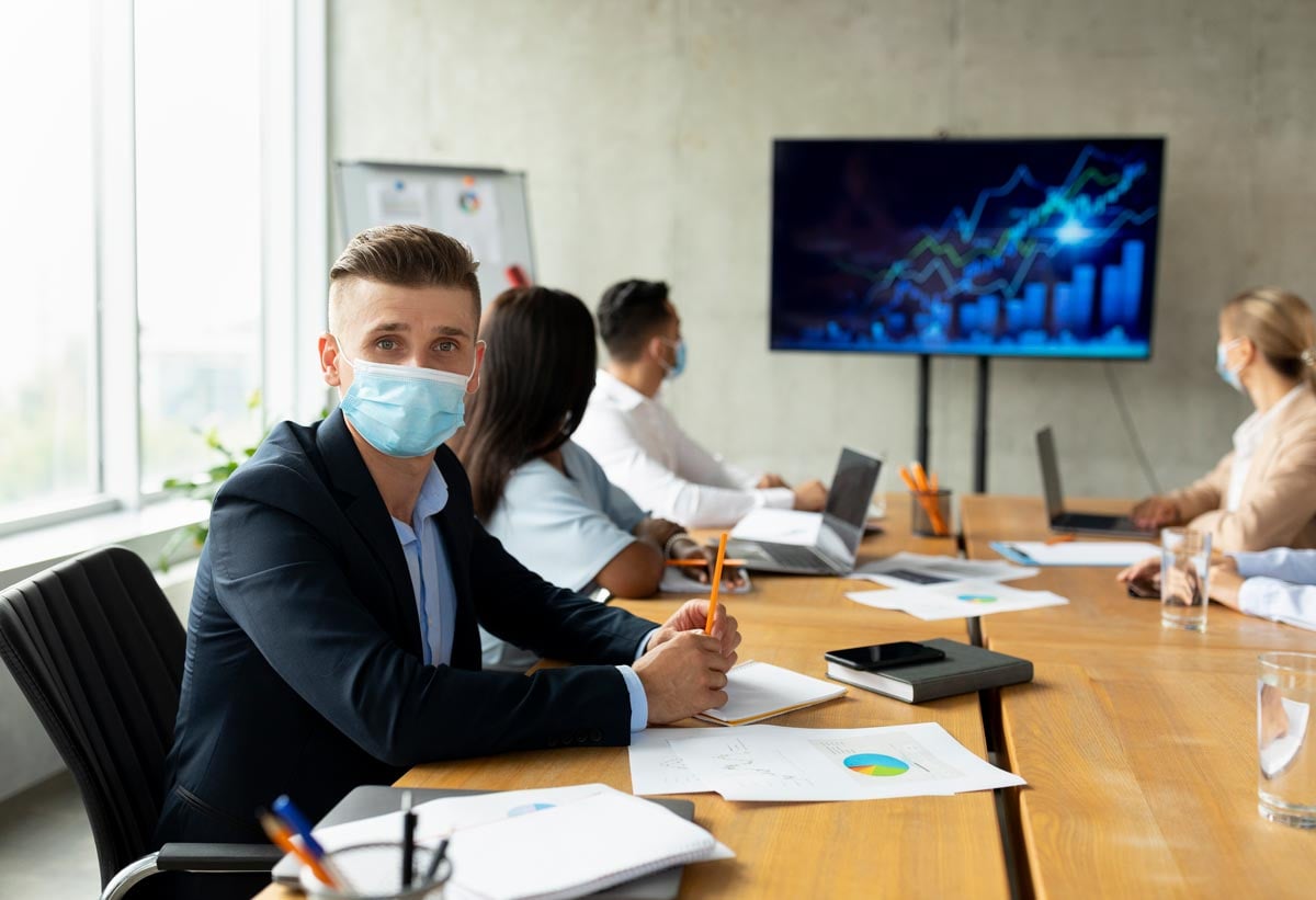 employee wearing a face mask sitting in a conference room with other colleagues looking at a presentation with a line chart